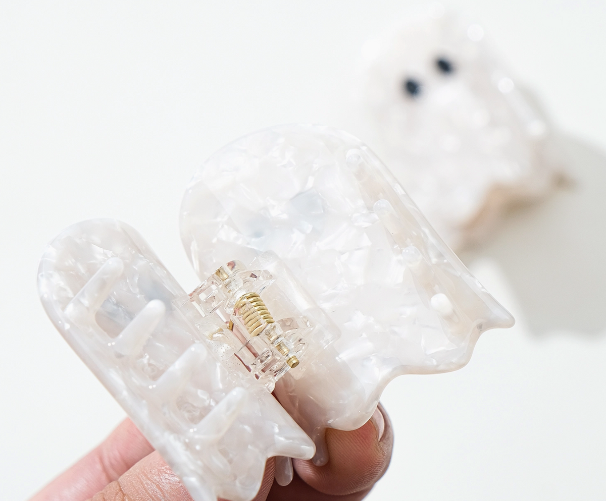 A translucent ghost-shaped hair claw clip being held by fingers against a clean white background, with a second blurred clip visible in the background.