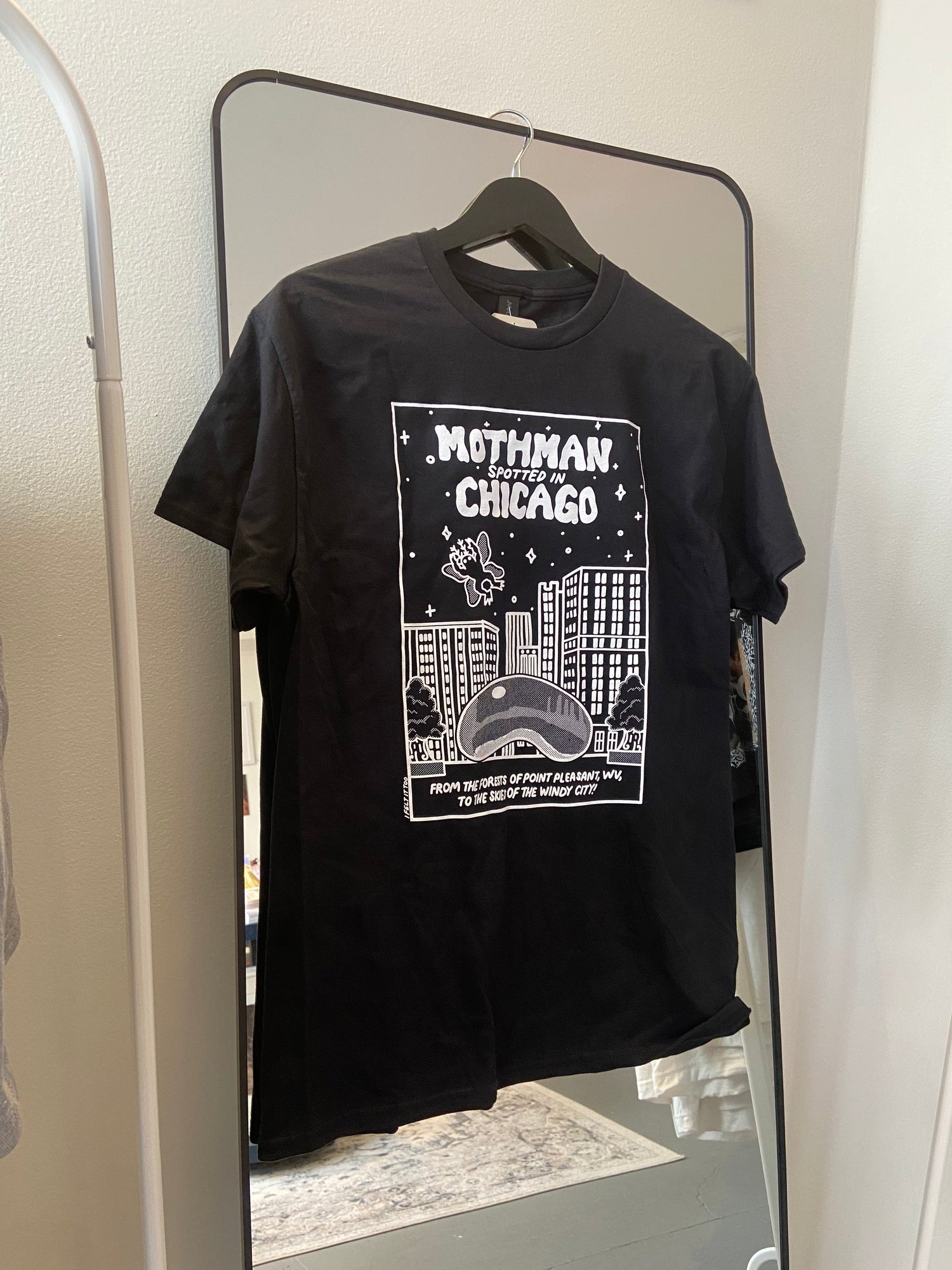Black t-shirt with graphic design hanging on a mirror. Design features Chicago's bean, and beloved cryptid Mothman in the sky.