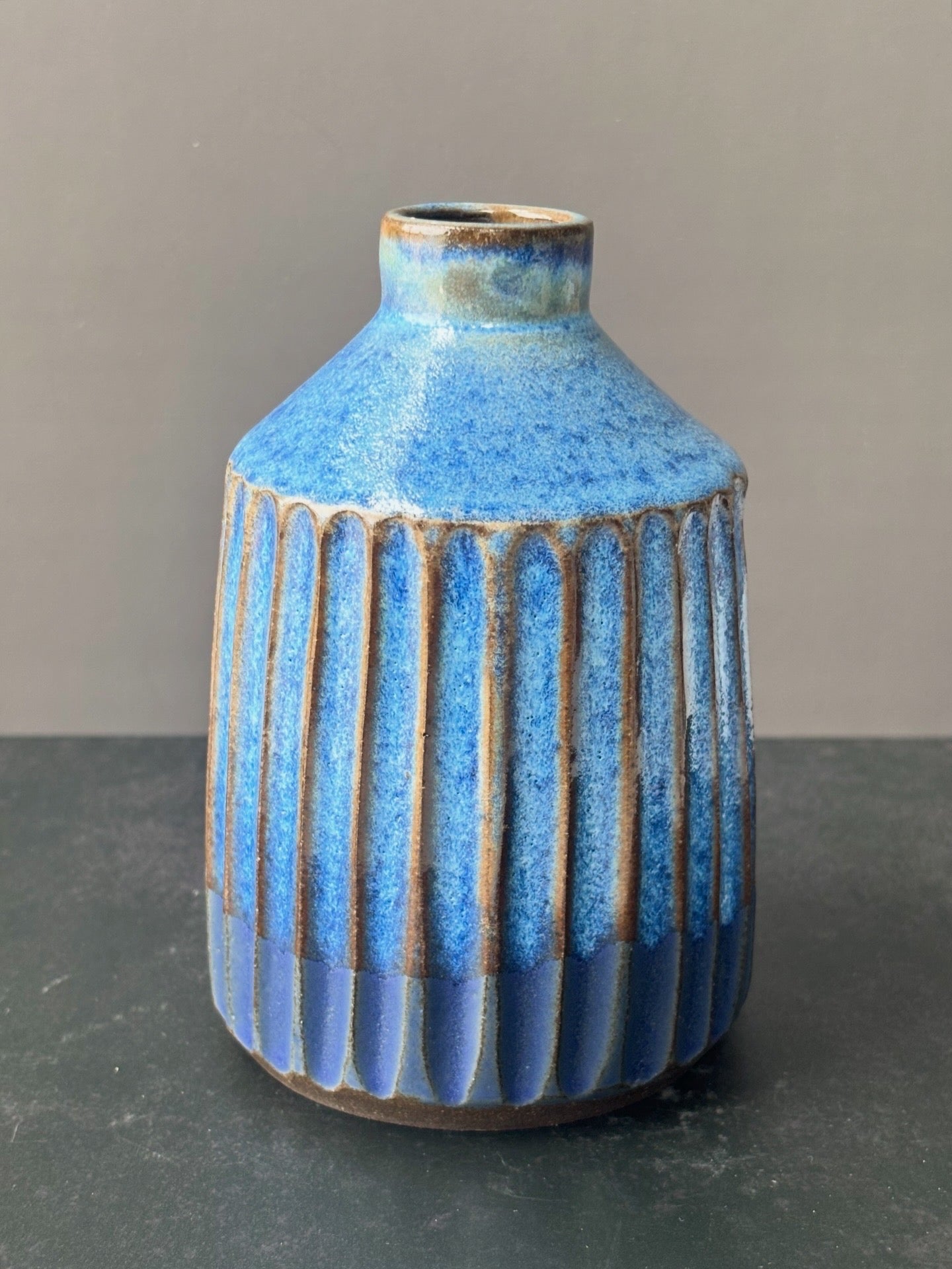 A small ceramic vase with a textured blue glaze, featuring vertical ribbed fluting on the lower half and a narrow neck, set against a neutral gray background.