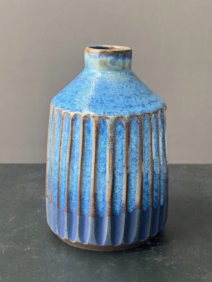 A small ceramic vase with a textured blue glaze, featuring vertical ribbed fluting on the lower half and a narrow neck, set against a neutral gray background.