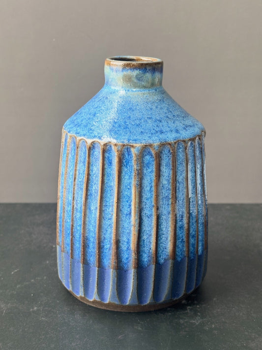 A small ceramic vase with a textured blue glaze, featuring vertical ribbed fluting on the lower half and a narrow neck, set against a neutral gray background.