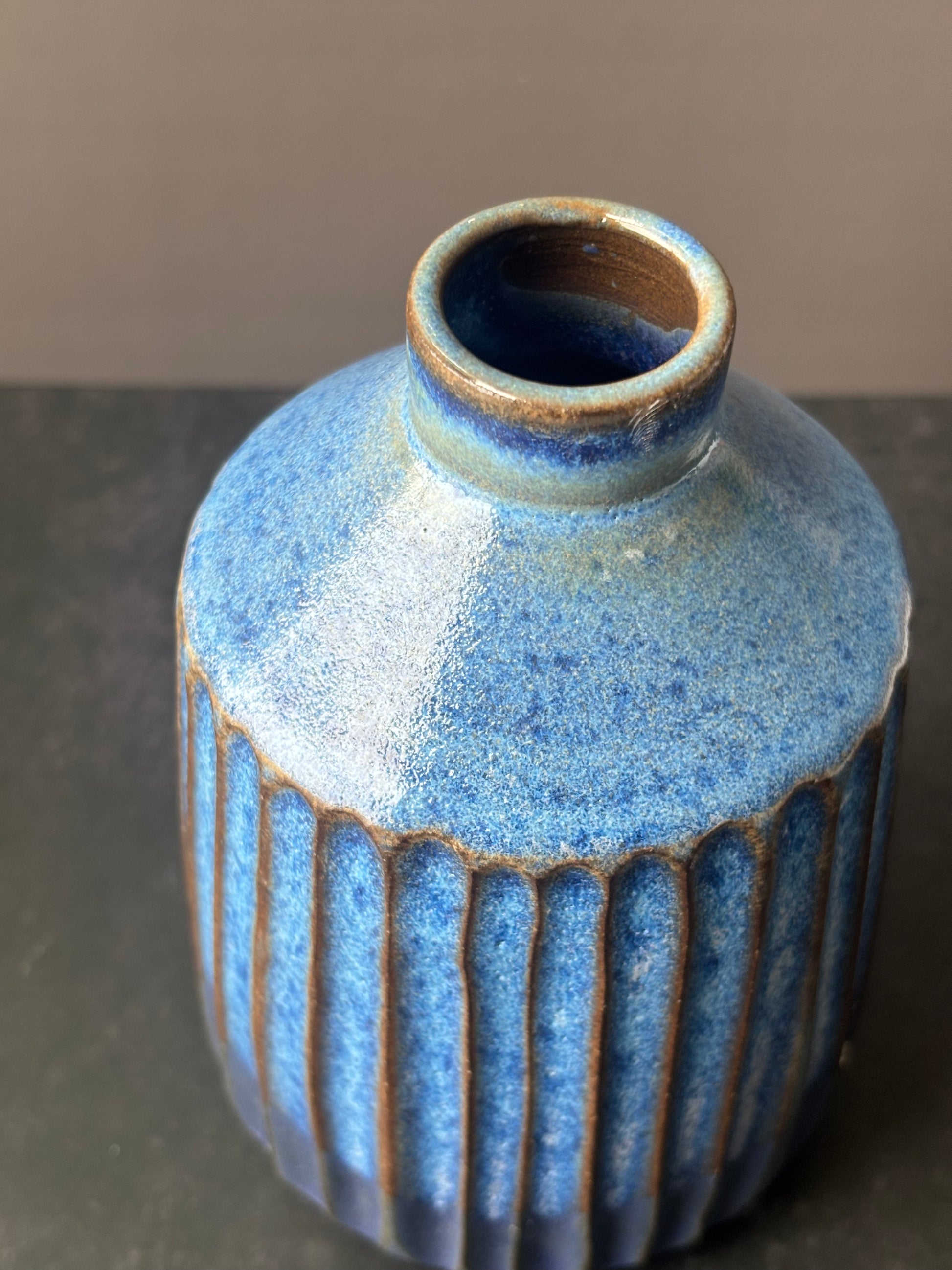 A small ceramic vase with a rounded body, flared neck, and vertical ribbed texture, covered in a speckled blue glaze with brown exposed clay at the rim and between the ribs, set against a neutral gray background.