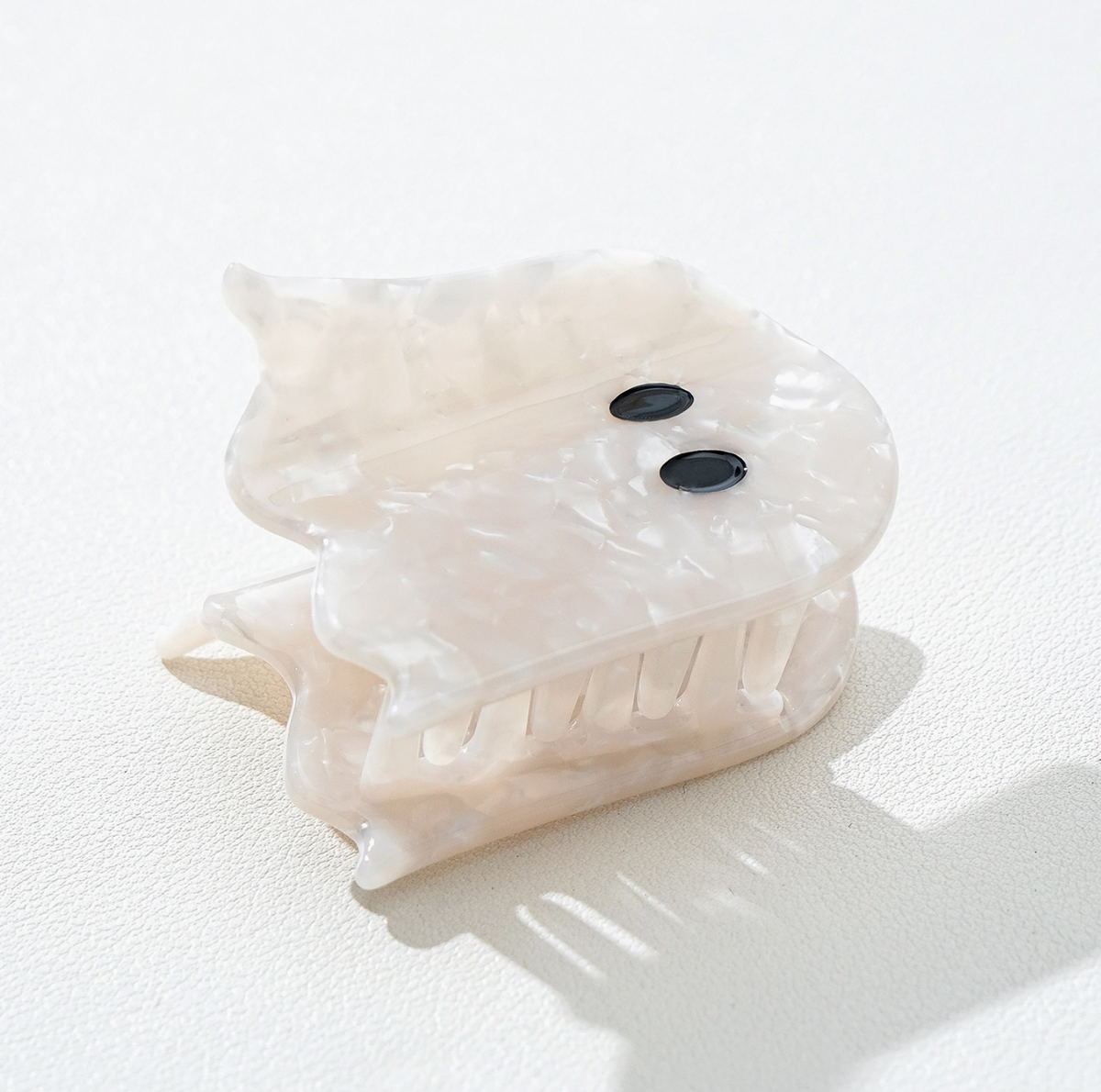 A translucent cream-colored ghost shaped hair claw clip. It has a textured, marbled finish and two small black oval eyes.  Photographed on a clean white surface with soft shadows.