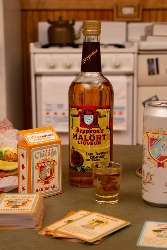 A bottle of Jeppson's Malört Liqueur stands on a kitchen counter next to a small filled shot glass, a can of beer, and the drinking game cards on a tabletop in a real kitchen setting.